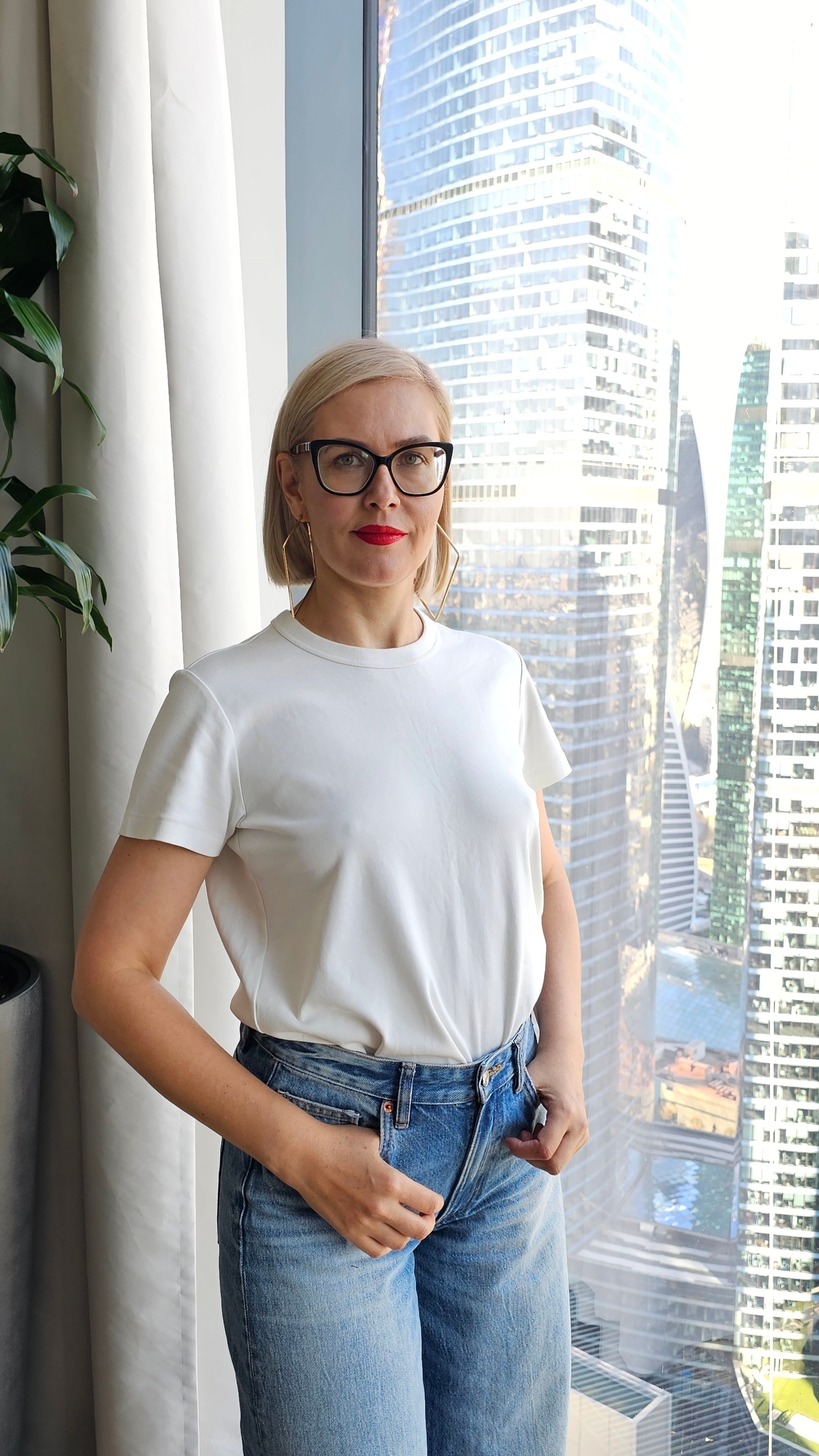 Девушка в белой футболке и джинсах стоит у окна