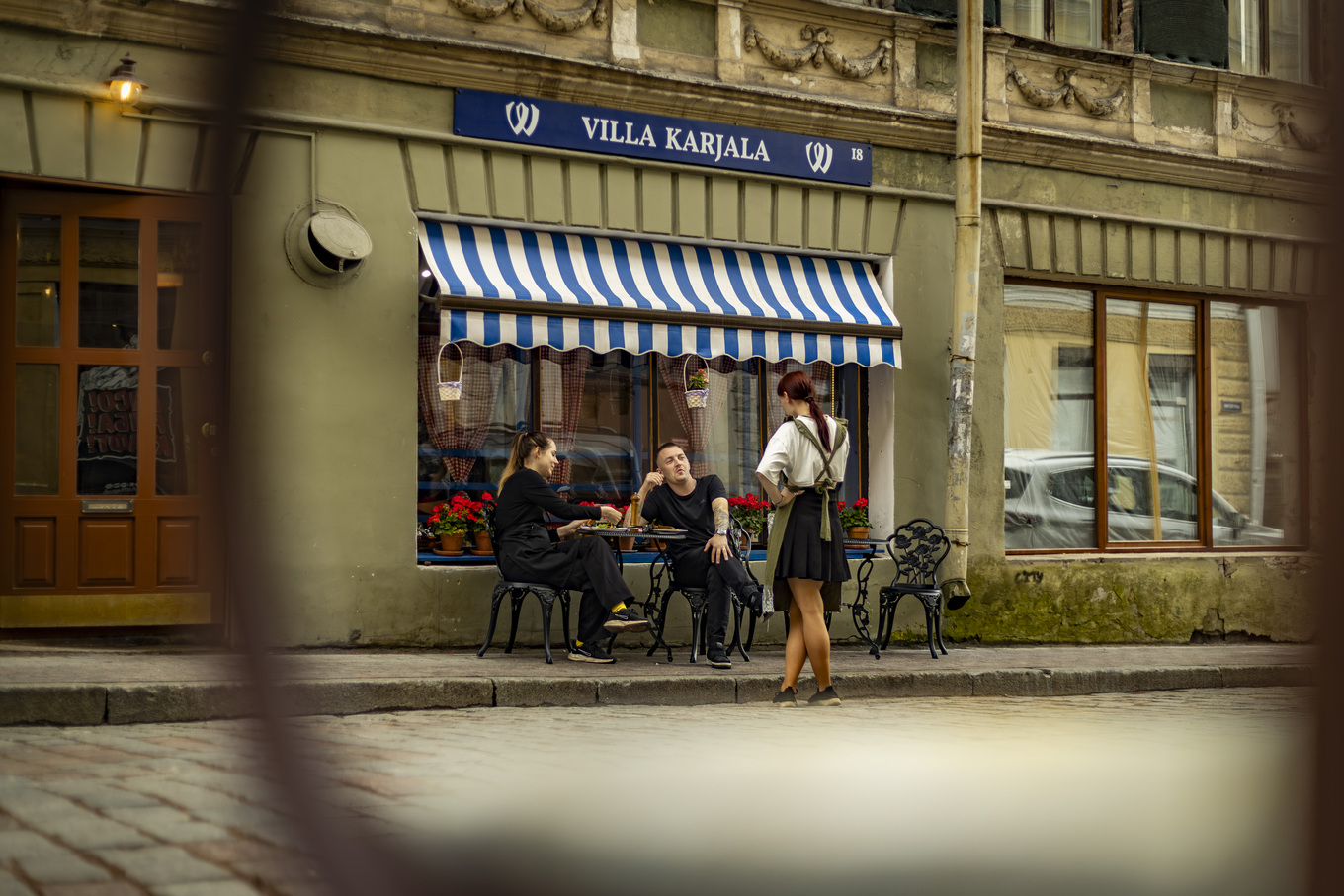 Фотография фасада кафе 'Villa Karjala' с сине-белым навесом; за столиком на улице сидят женщина и мужчина, а рядом стоит девушка в фартуке, возможно, официантка; фон — старинное здание и улица