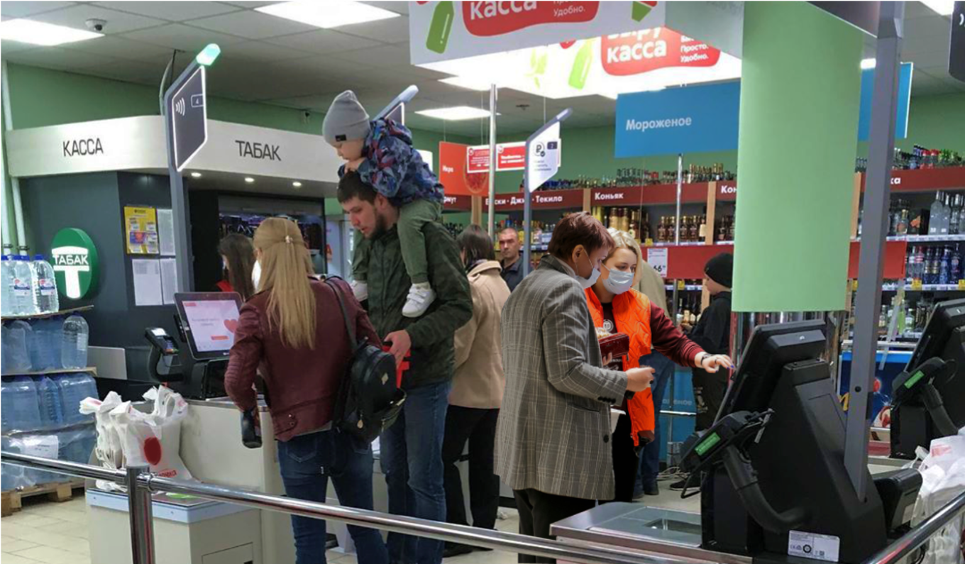 Очередь в супермаркете: покупатели с детьми, тележками и сумками стоят у касс, один из сотрудников в маске и жилете помогает на линии