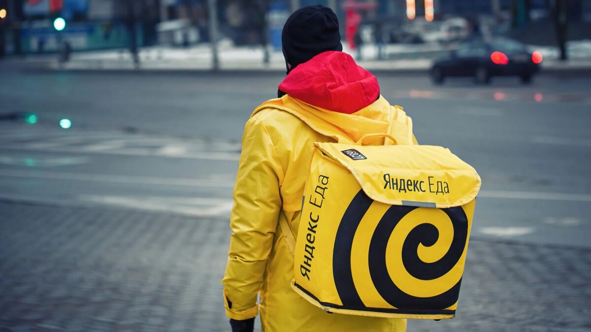 Курьер Яндекс еды в желтой куртке с желтым фирменным рюкзаком идёт по улице