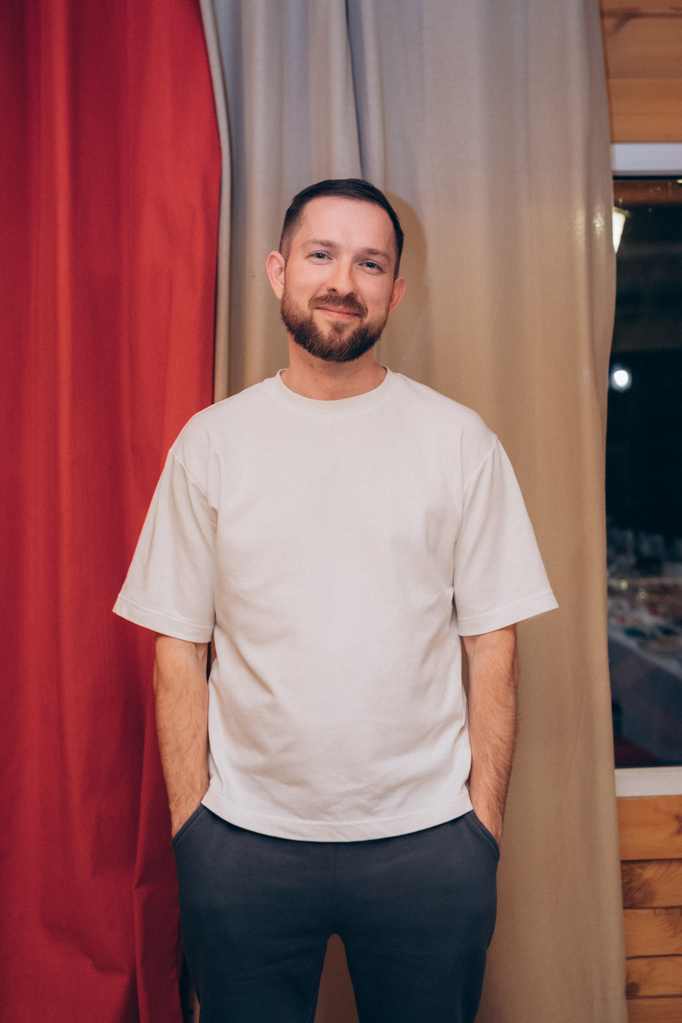 Руслан Гатиятов в белой футболке и темных брюках стоит на фоне красной и белой ткани, улыбаясь