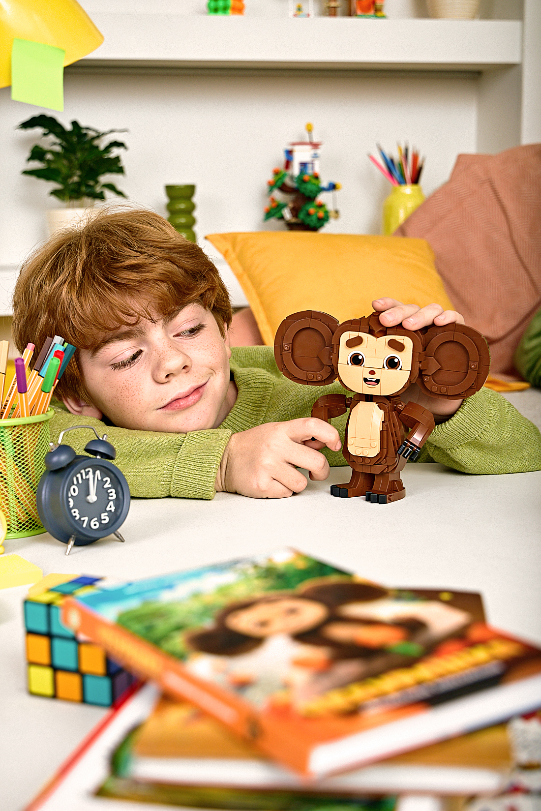 Рыжеволосый мальчик с веснушками в зеленом свитере сидит за столом и с улыбкой собирает фигурку Чебурашки из конструктора