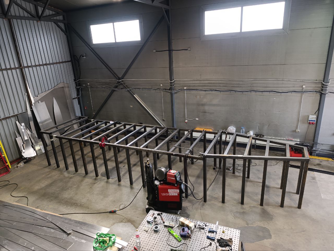 Промышленный цех сварочного производства. Длинный металлический каркас на полу, рядом сварочный аппарат, стол с инструментами