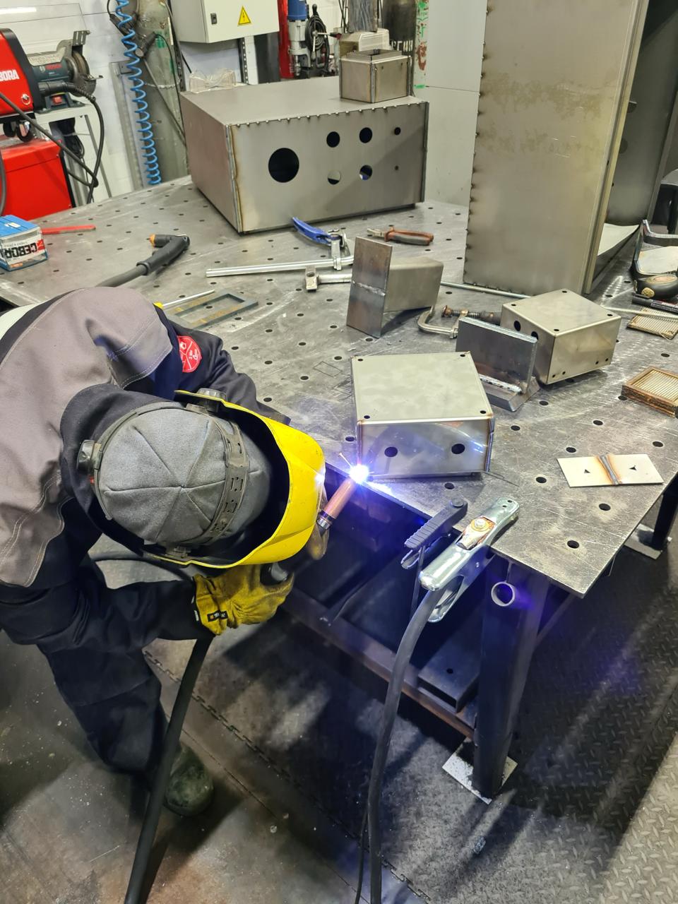 Сварщик в защитной маске и перчатках варит TIG шов на металлическом коробе на сборочном столе, вокруг заготовки и инструменты