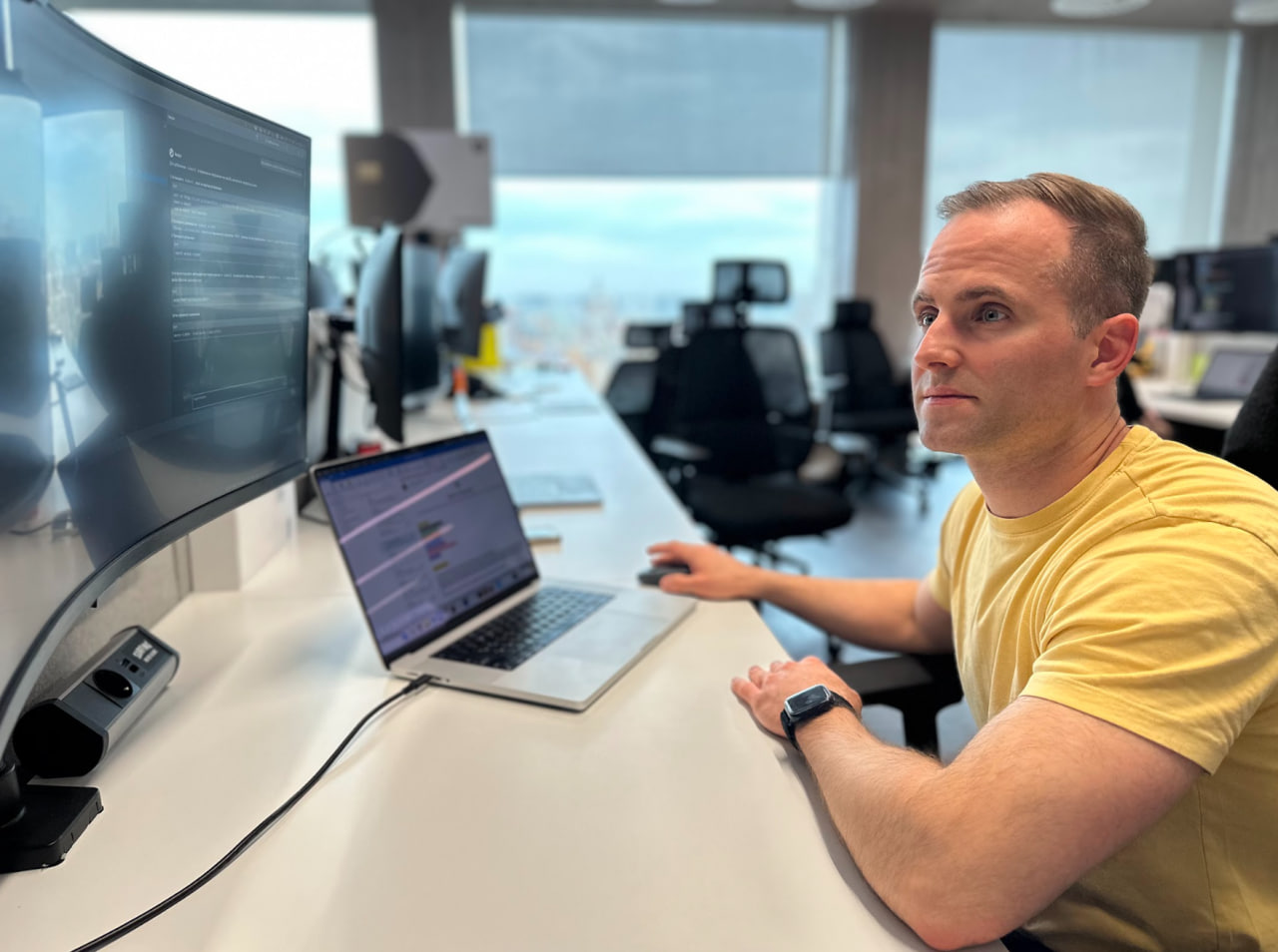 Антон Чудин, backend-разработчик работает за ноутбуком и следит за метриками на широкоформатном мониторе в современном офисе с панорамными окнами