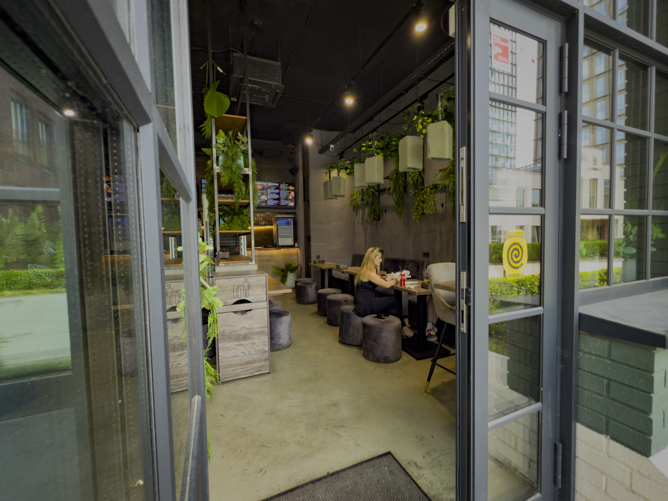 Вход в современное кафе с растениями, мягкими пуфами и посетителями за столиками
