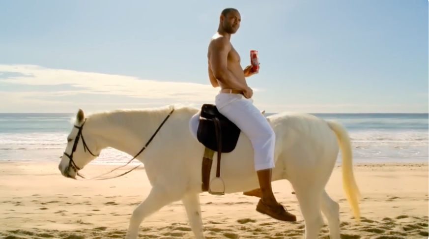 Мужчина верхом на белой лошади идёт по пляжу, держит в руке банку газировки и смотрит в сторону