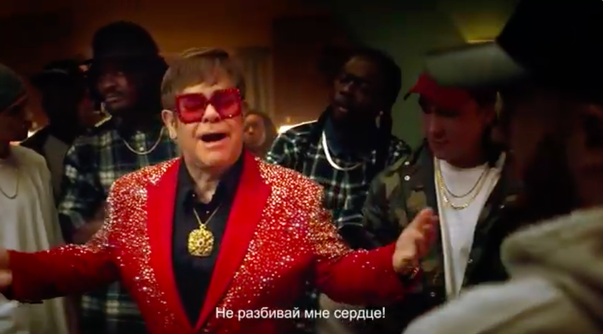 Элтон Джон в красном блестящем пиджаке и больших очках поёт перед группой мужчин в уличной одежде