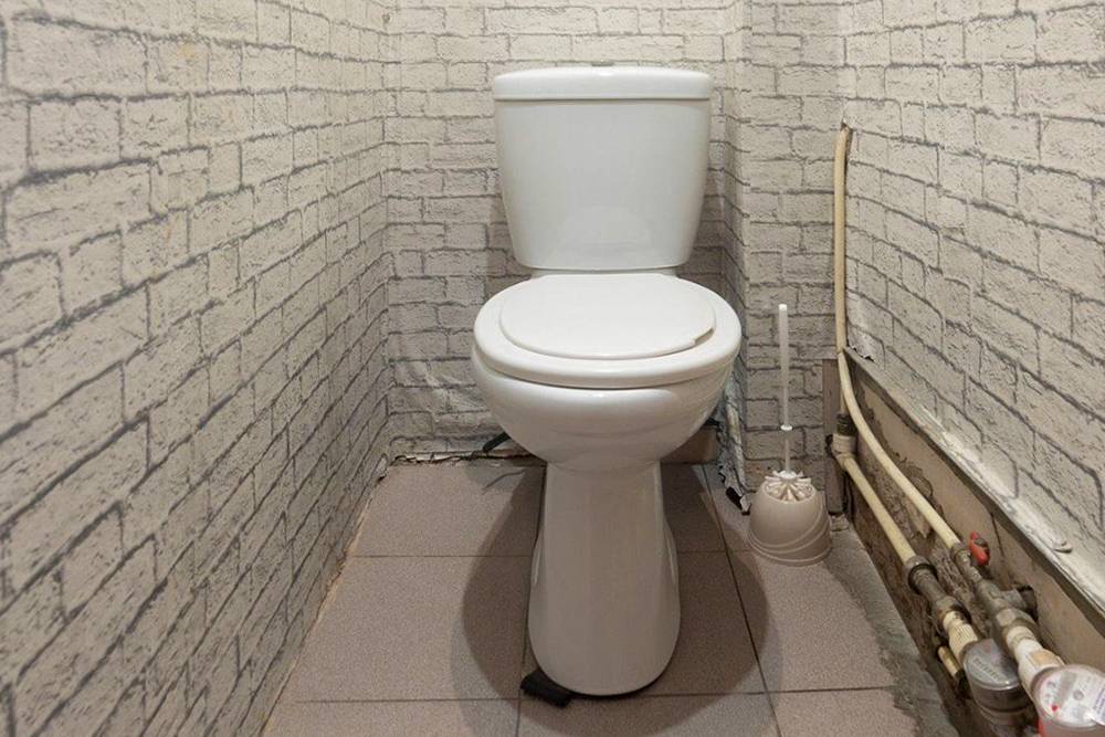 Туалет до ремонта. На стенах обои, на полу плитка. Трубы вдоль стены и счетчики не спрятаны