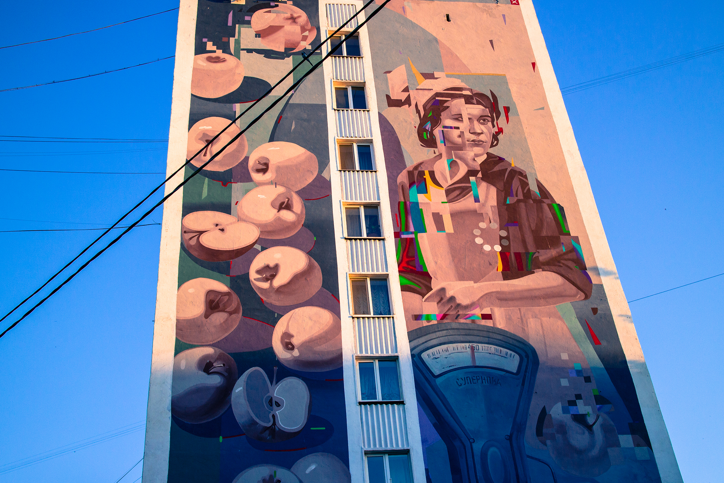 Этот мурал, как и подавляющее большинство, нарисовали участники фестиваля уличного искусства «Меморандум», который проходил в Уфе с 2022 по 2024 год. Конкретно к этому арт-проекту местные относятся неоднозначно. Досталось и своеобразному изображению лица, стилизованному под глитчГлитч — направление в изобразительном искусстве, в котором цифровые ошибки и помехи используют как эстетический инструмент самовыражения, и иконке загрузки. Здание расположено в районе рынка, и, как я понял задумку автора, мурал через соответствующую отсылку призван показать связь прошлого с настоящим