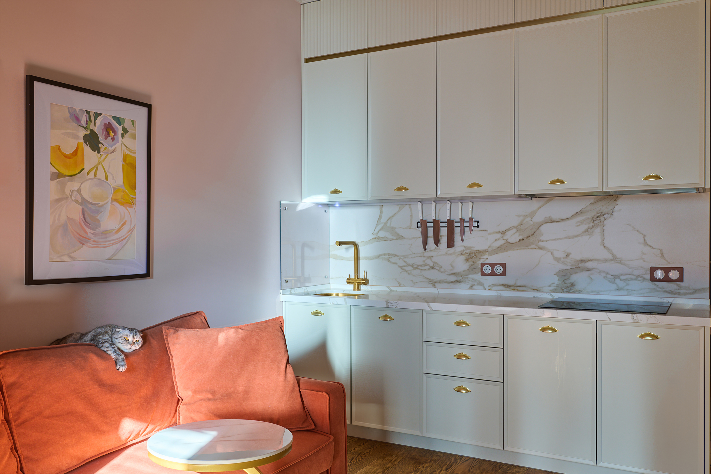 Золотые ручки выглядят нестандартно и сочетаются со смесителем и такой же линией-вставкой над фасадами. Картина дополняет нежную палитру
