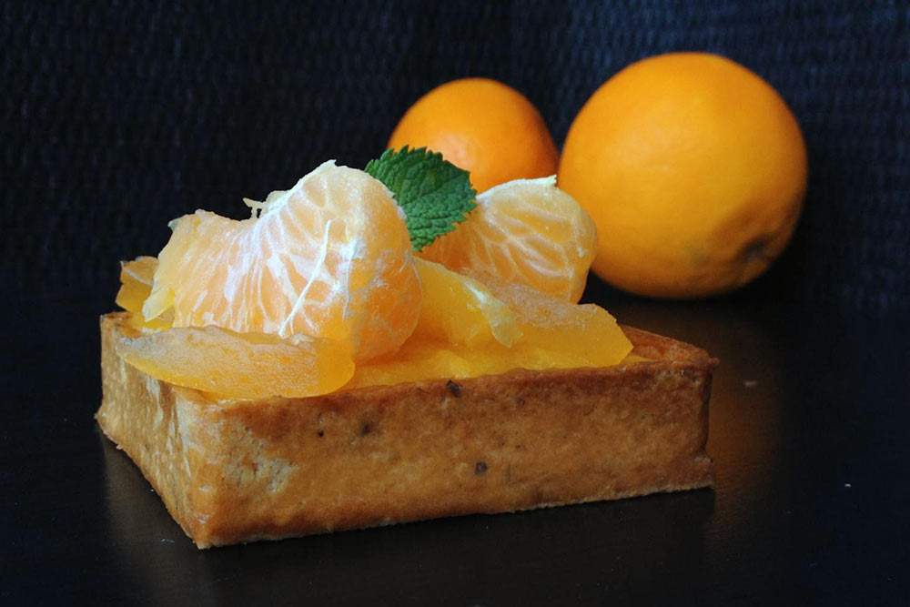 Апельсиновая тарталетка, выпеченная в квадратной перфорированной форме. Состав: песочная основа, миндальный крем с апельсином, кремю из апельсина с ликером «Куантро», мармелад из мандаринов. Украшена дольками свежих апельсинов, мандаринов, апельсиновыми цукатами собственного изготовления и листиком мяты