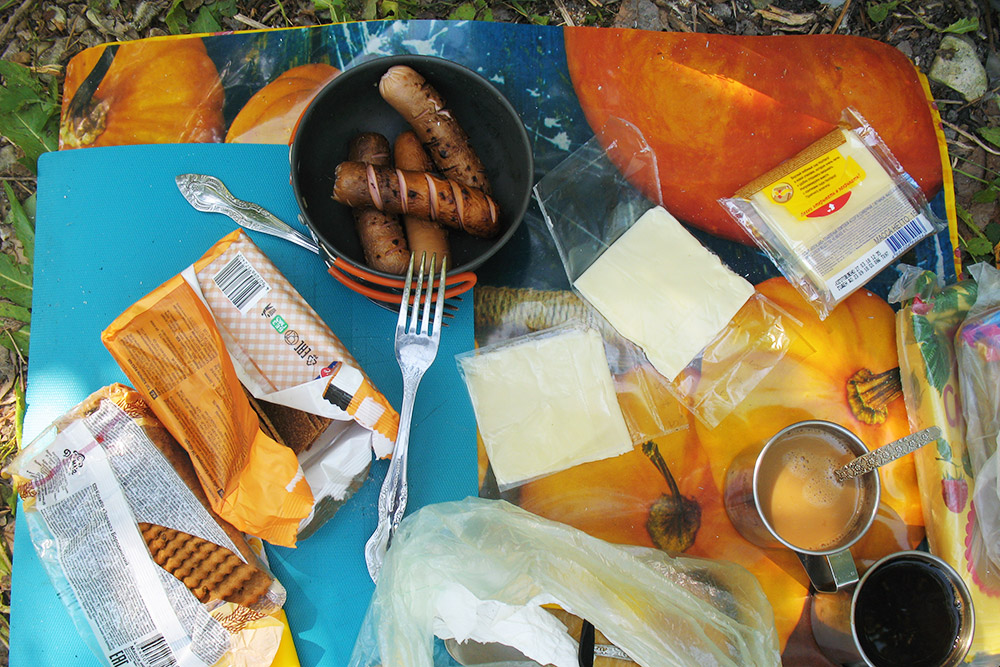 Так выглядит походный обед: клеенка на траве, железные кружки с кофе, крекер, хрустящие хлебцы с плавленым сыром и жареные сосиски. Шпажки для сосисок мы делали из ивовых веток
