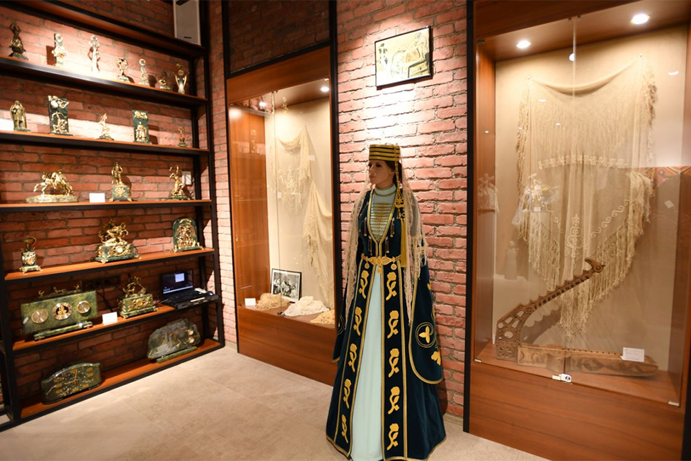 На манекене представлен традиционный костюм, за ним — старинные платки, вышитые вручную, на полке находится музыкальный инструмент — двенадцатиструнная лира. Источник: alania.gov.ru