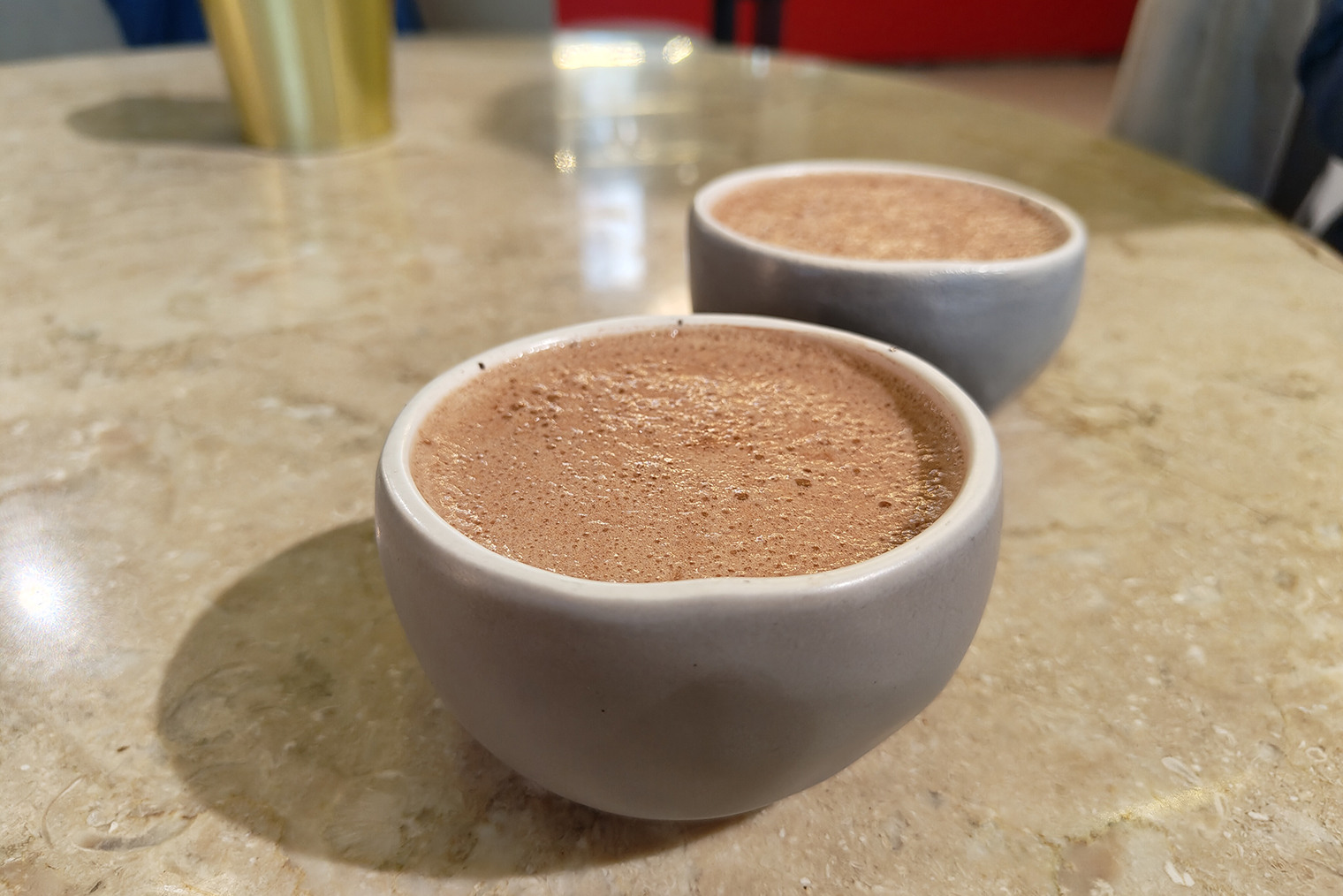 Кофейный напиток «брусника⁠-⁠батч» по виду больше напоминает какао. Камера сохранила ореховый оттенок пенки и текстуру с множеством пузырьков. Задний фон при этом аккуратно размыт