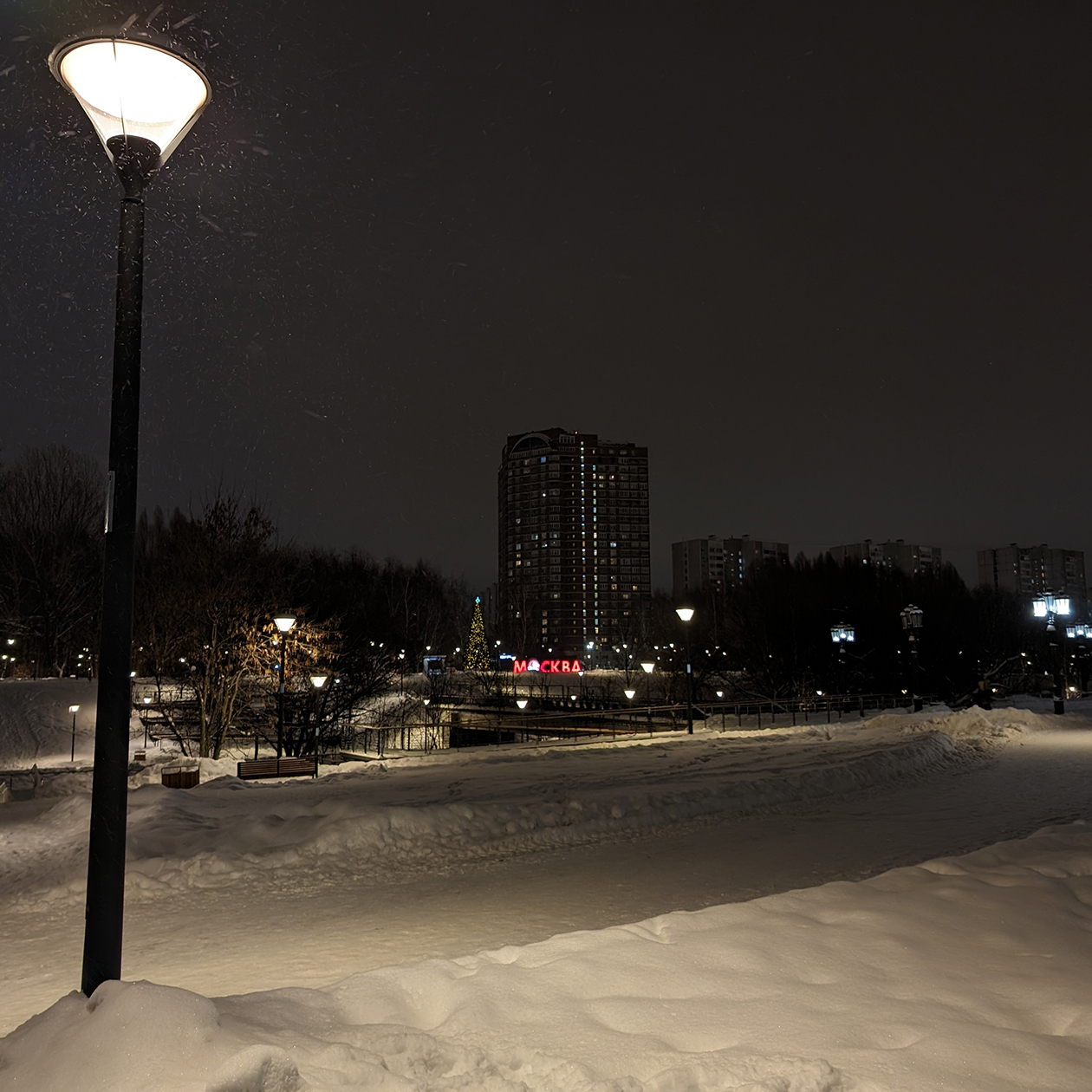 Ночью та же сцена выглядит совсем иначе: фонари немного засвечены, а четкость домов вдали ощутимо упала, но вот снег в свете фонаря смотрится даже более фактурным и проработанным, чем днем